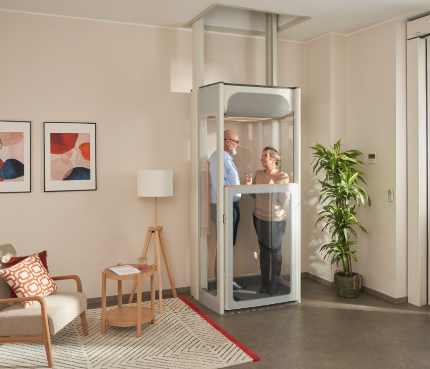 Et eldre par står i en Stannah Uplifts-boligheis i en lys stue, med et kompakt og gjennomsiktig gulvdesign og klare paneler.